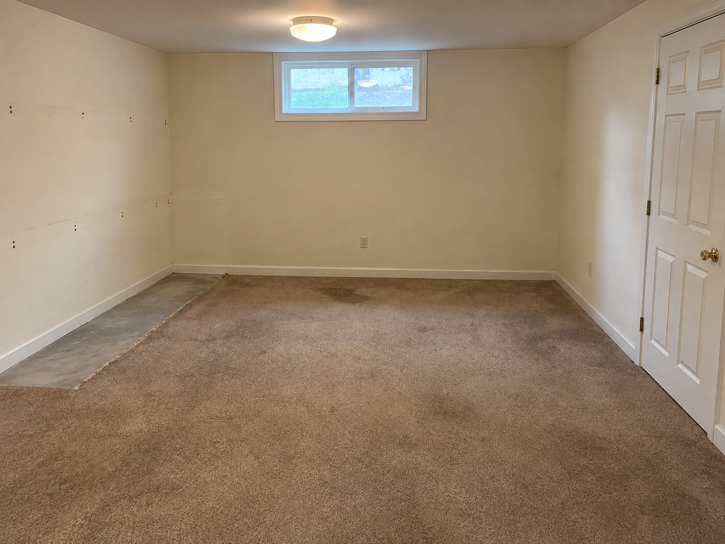 Basement after removal of fixed cabinets and wall shelf, with existing worn carpet retained and cabinet footprint exposed.