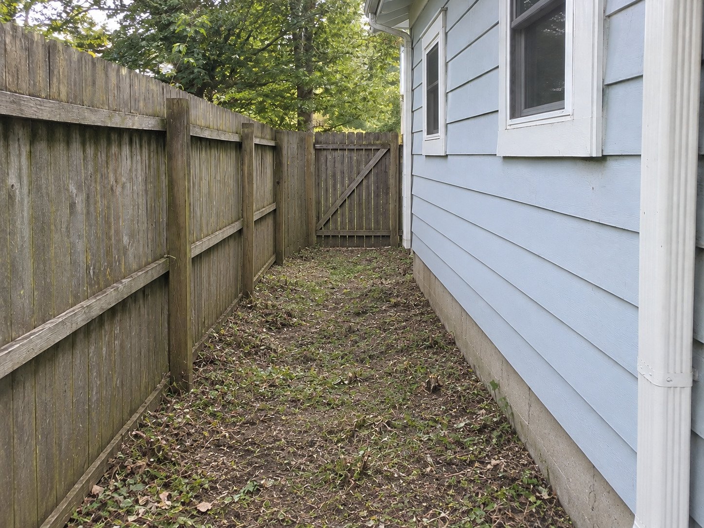 Side access reopened after brush removal, with the old soil and retained fence and house unchanged.