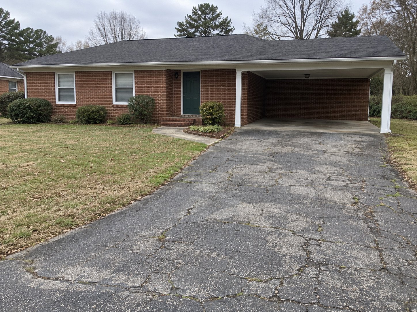 Cracked asphalt driveway leading to a brick home's empty carport before paving.