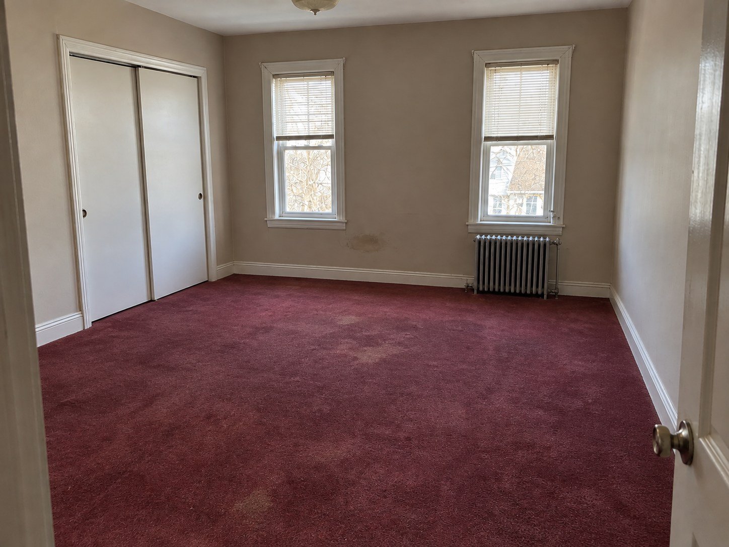 The same rental bedroom cleared with original worn carpet, wall scuff and radiator retained.
