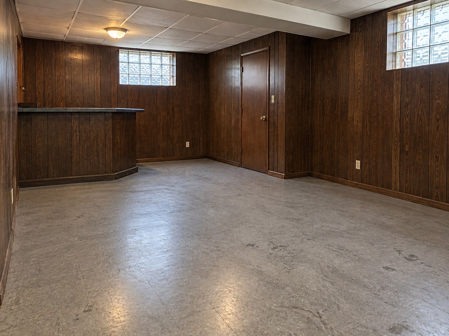 The same basement cleared of movable contents while retaining its built-in bar and worn vinyl floor.