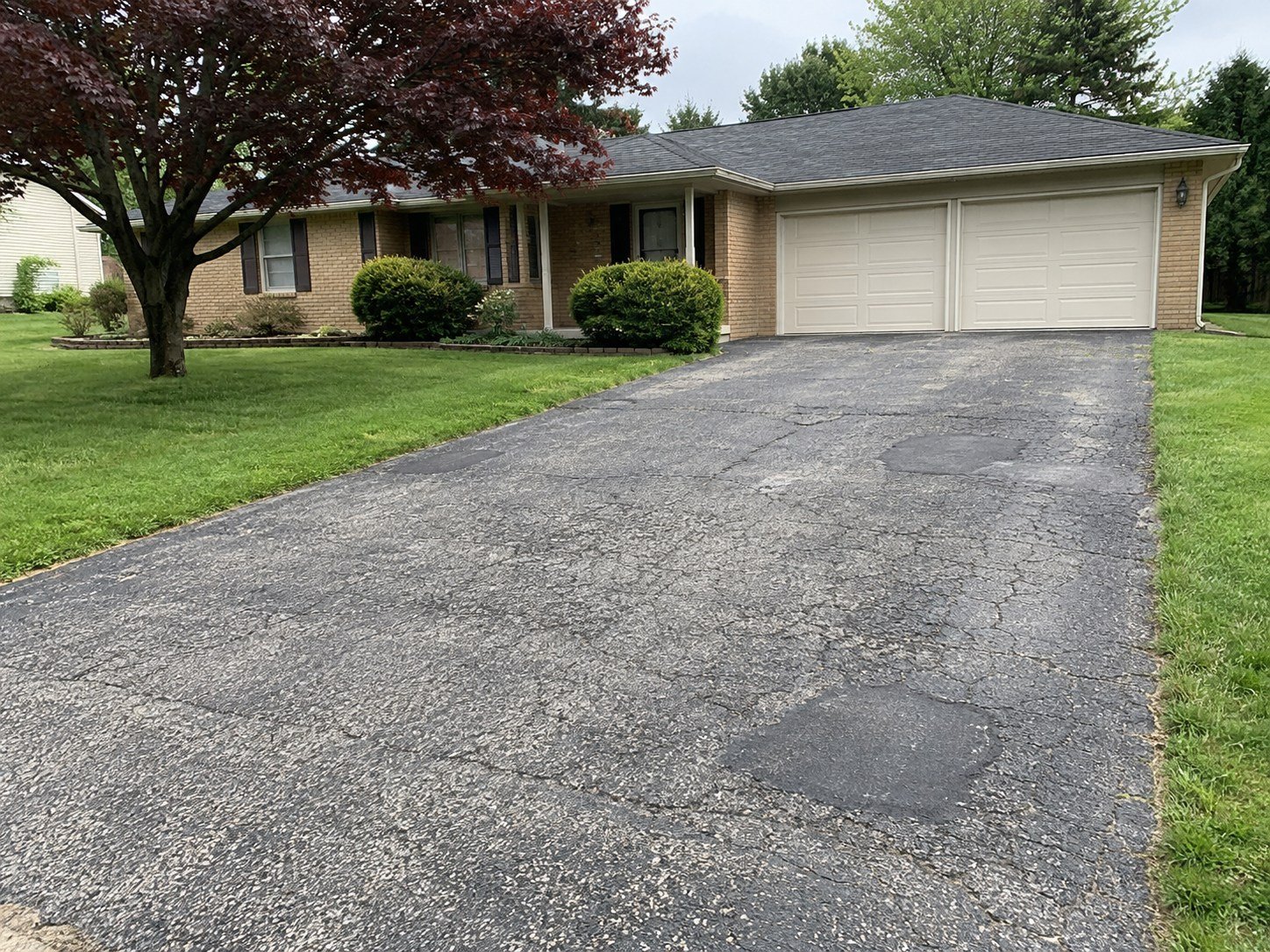 A worn cracked asphalt driveway in front of a tan brick ranch and two-car garage.