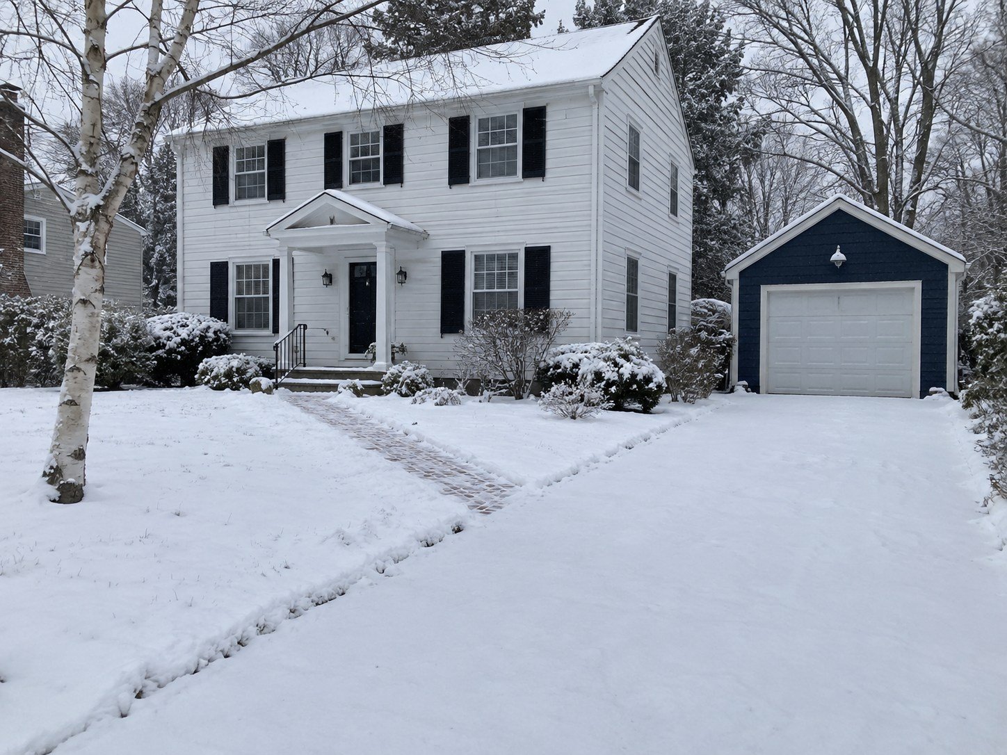 Snow-covered driveway beside a white Colonial house and navy detached garage.
