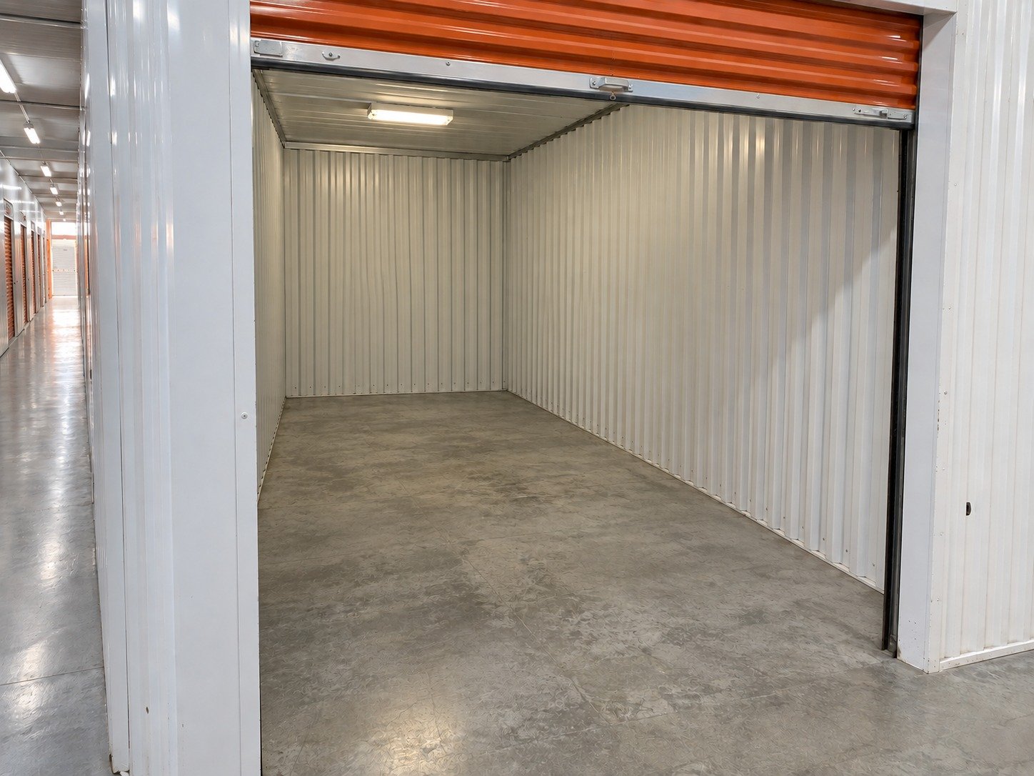 The same indoor storage locker emptied, retaining its orange shutter, white partitions and worn concrete floor.