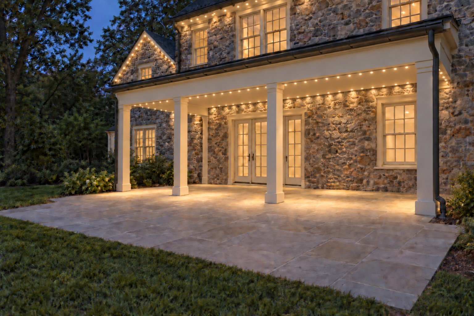 Stone-home covered patio with warm-white lighting beneath the porch perimeter, upper house eave and visible gable edges.