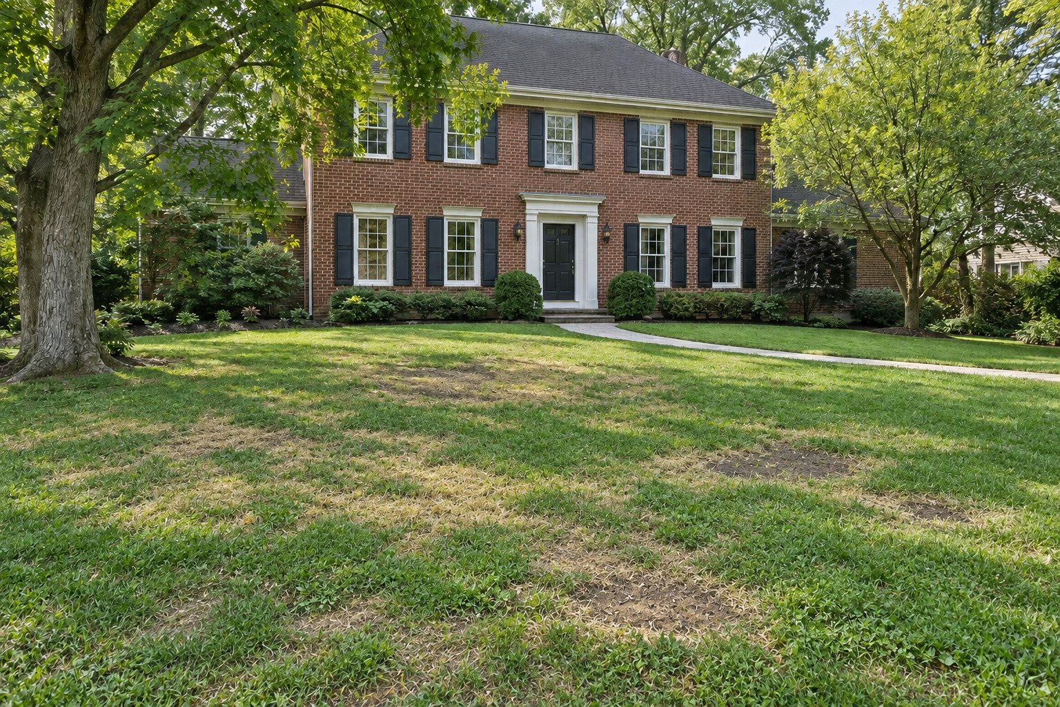 Patchy front lawn with thin grass and worn soil areas at an established brick Georgian home before turf restoration.