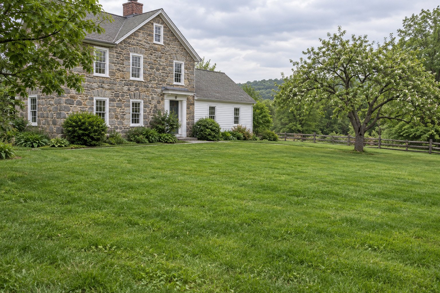 Restored green farmhouse lawn on the same gentle grade, with the stone home, mature tree, beds and boundary fence retained.